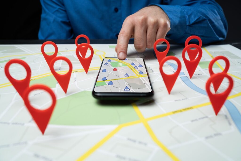 Eine Hand tippt auf ein Smartphone, das auf einer gedruckten Landkarte liegt. Auf dem Bildschirm und der Karte sind zahlreiche rote Standort-Markierungen zu sehen. Die Szene symbolisiert lokale Suchmaschinenoptimierung (Local SEO), Standortsuche oder digitale Karten- und Navigationsdienste.