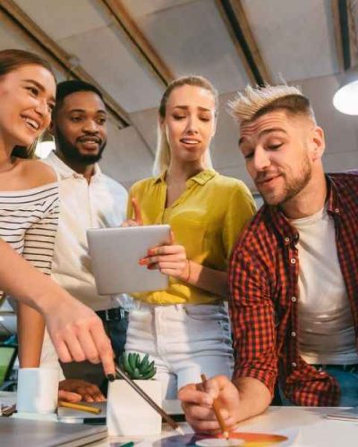 Eine Gruppe junger Menschen arbeitet gemeinsam an einem kreativen Projekt in einem modernen Büro. Sie diskutieren, zeigen auf Farbmuster, nutzen ein Tablet und wirken engagiert. Das Bild symbolisiert Teamarbeit, Designprozesse oder kreative Agenturarbeit.